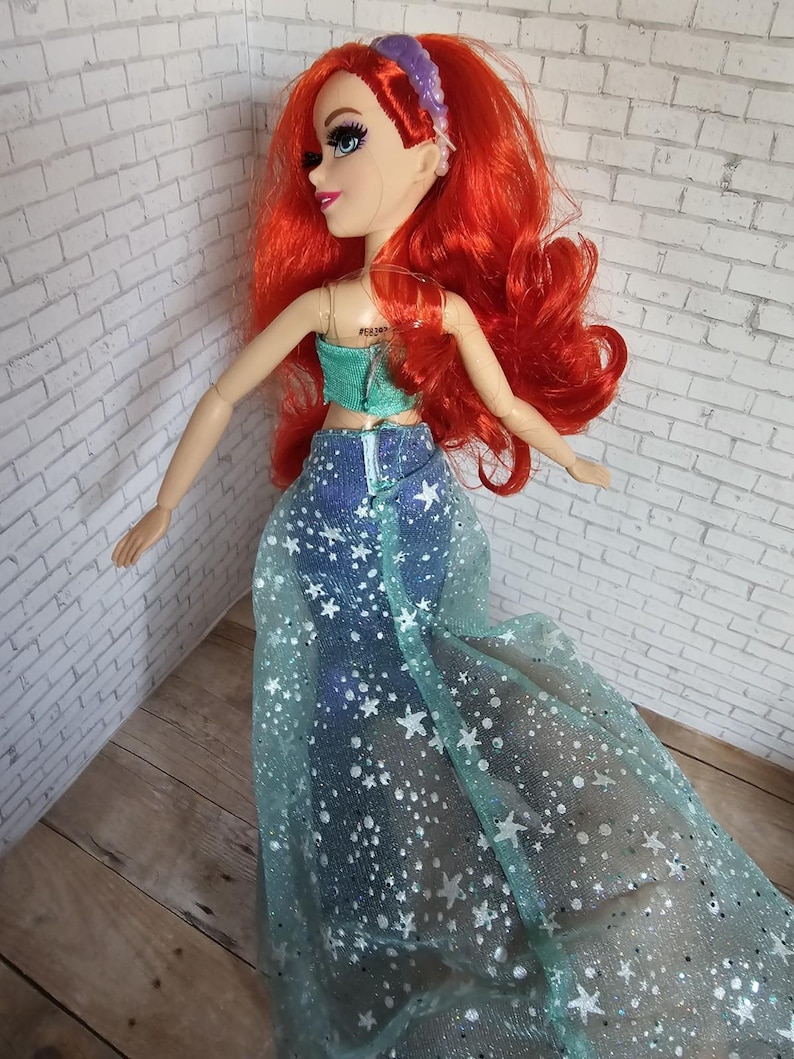 May include: A doll with bright red hair, wearing a teal top and a denim skirt with a sheer, star-patterned overlay. The doll has blue eyes and a purple headband. The doll is posed against a white brick wall.