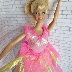 Pode incluir: Uma boneca loira com cabelo preso num rabo de cavalo alto, vestindo um vestido rosa e dourado. O corpete apresenta um tecido rosa metálico cintilante com folhos, enquanto a saia consiste em camadas de tule rosa e tecido dourado metálico.