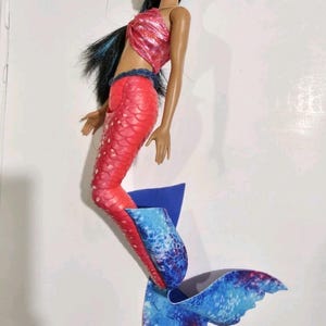 Puede incluir: Mu&ntilde;eca de sirena con top rosa con lentejuelas y cola roja con estampado de escamas. La cola de la mu&ntilde;eca hace la transici&oacute;n a una aleta azul y blanca estampada. La mu&ntilde;eca tiene cabello negro y est&aacute; posando como si nadara.
