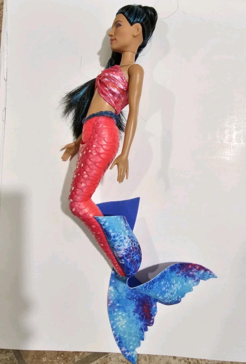 Puede incluir: Mu&ntilde;eca de sirena con top rosa con lentejuelas y cola roja con estampado de escamas. La cola de la mu&ntilde;eca hace la transici&oacute;n a una aleta azul y blanca estampada. La mu&ntilde;eca tiene cabello negro y est&aacute; posando como si nadara.