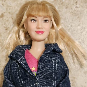 Op de afbeelding: Een pop met blond haar en een spijkerjasje over een roze top. De pop heeft een glimlachende uitdrukking met roze lippenstift. De jas heeft witte stiksels. Het haar van de pop is gestyled met pony en staartjes.