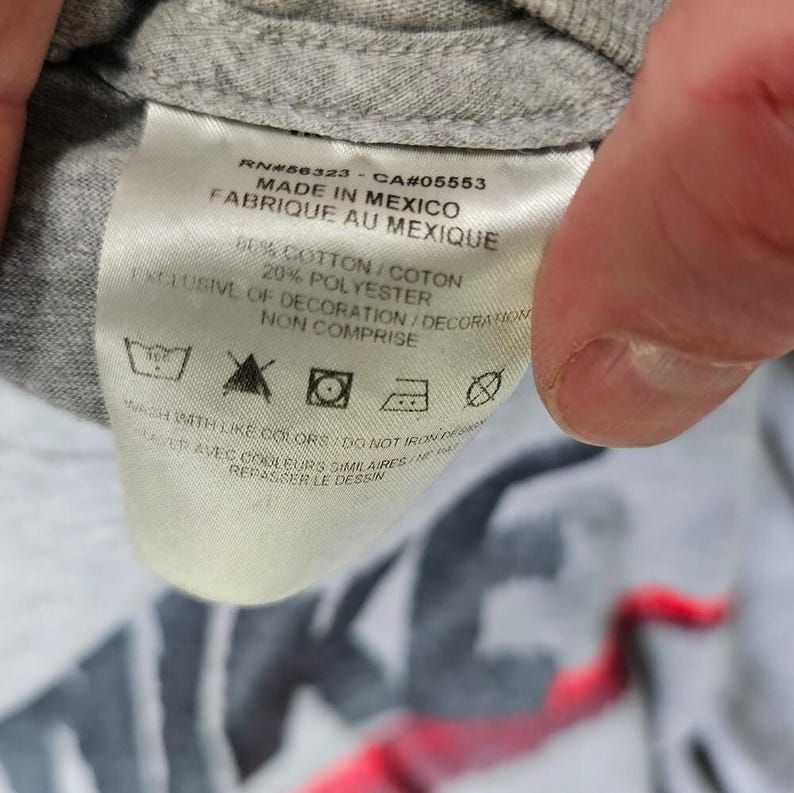 Pu&ograve; includere: Primo piano di un'etichetta di una maglietta grigia con testo. L'etichetta recita "MADE IN MEXICO" ed elenca il contenuto del tessuto come 80% cotone e 20% poliestere. Le istruzioni di lavaggio sono anch'esse stampate.