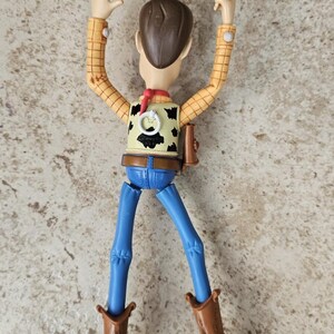 May include: A Woody action figure from the Toy Story franchise. The figure is wearing a yellow plaid shirt, a cow-print vest, blue jeans, and brown cowboy boots. The figure has brown hair and a red neckerchief. The figure is posed with arms raised.