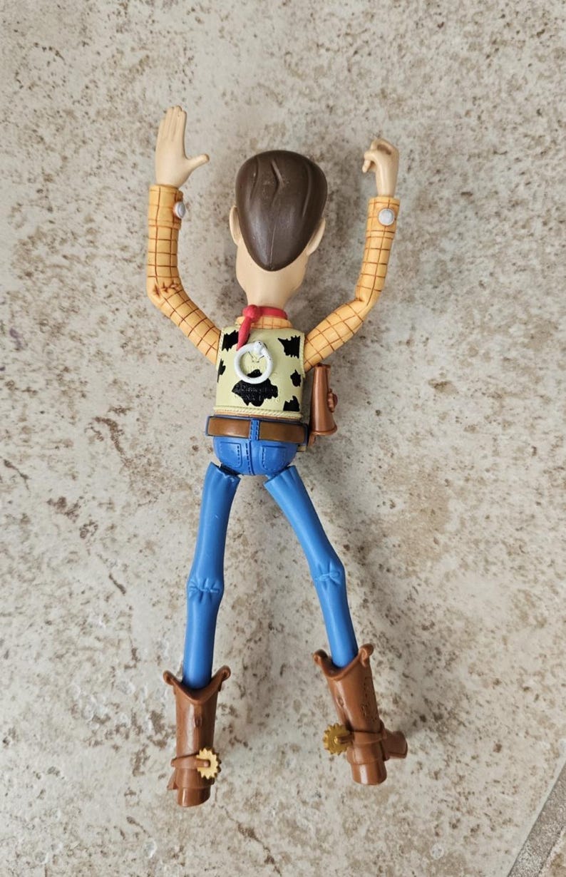 May include: A Woody action figure from the Toy Story franchise. The figure is wearing a yellow plaid shirt, a cow-print vest, blue jeans, and brown cowboy boots. The figure has brown hair and a red neckerchief. The figure is posed with arms raised.