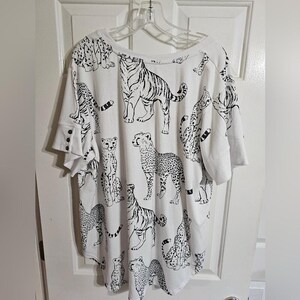 May include: White short-sleeved shirt with a black animal print featuring tigers, cheetahs, and other big cats. The shirt has a wide neckline and a relaxed fit. The sleeves have a button detail.