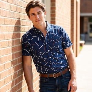May include: A navy blue short-sleeve button-down shirt with a white and red dash pattern. The shirt is paired with blue jeans and a brown leather belt. The person is leaning against a brick wall.