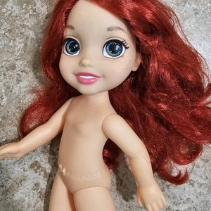 May include: A doll with red hair and a smiling face. The doll has large blue eyes and a light skin tone. The doll's arms are outstretched, and it is missing clothing. The doll is a character from a popular animated film.