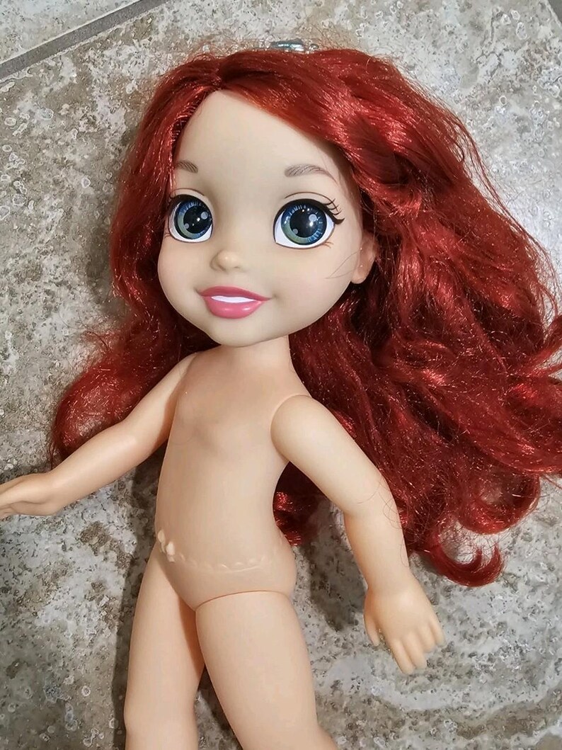 May include: A doll with red hair and a smiling face. The doll has large blue eyes and a light skin tone. The doll's arms are outstretched, and it is missing clothing. The doll is a character from a popular animated film.