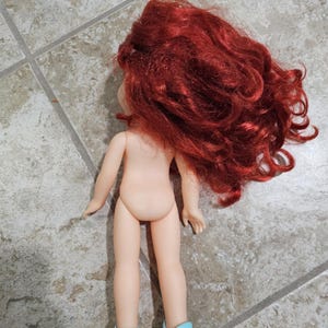 May include: A doll with red curly hair and light blue shoes. The doll is nude and facing away from the camera. The doll's hair is voluminous and covers the back of the doll's head and shoulders. The doll is standing on a tiled surface.