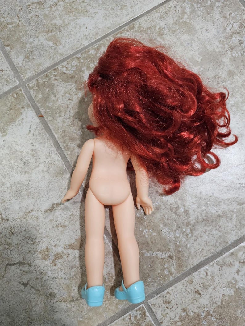 May include: A doll with red curly hair and light blue shoes. The doll is nude and facing away from the camera. The doll's hair is voluminous and covers the back of the doll's head and shoulders. The doll is standing on a tiled surface.