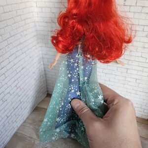 May include: A doll with bright red hair and a blue dress decorated with white stars. The doll's dress has a sheer, light blue overlay with white star accents. The doll is being held by a person.