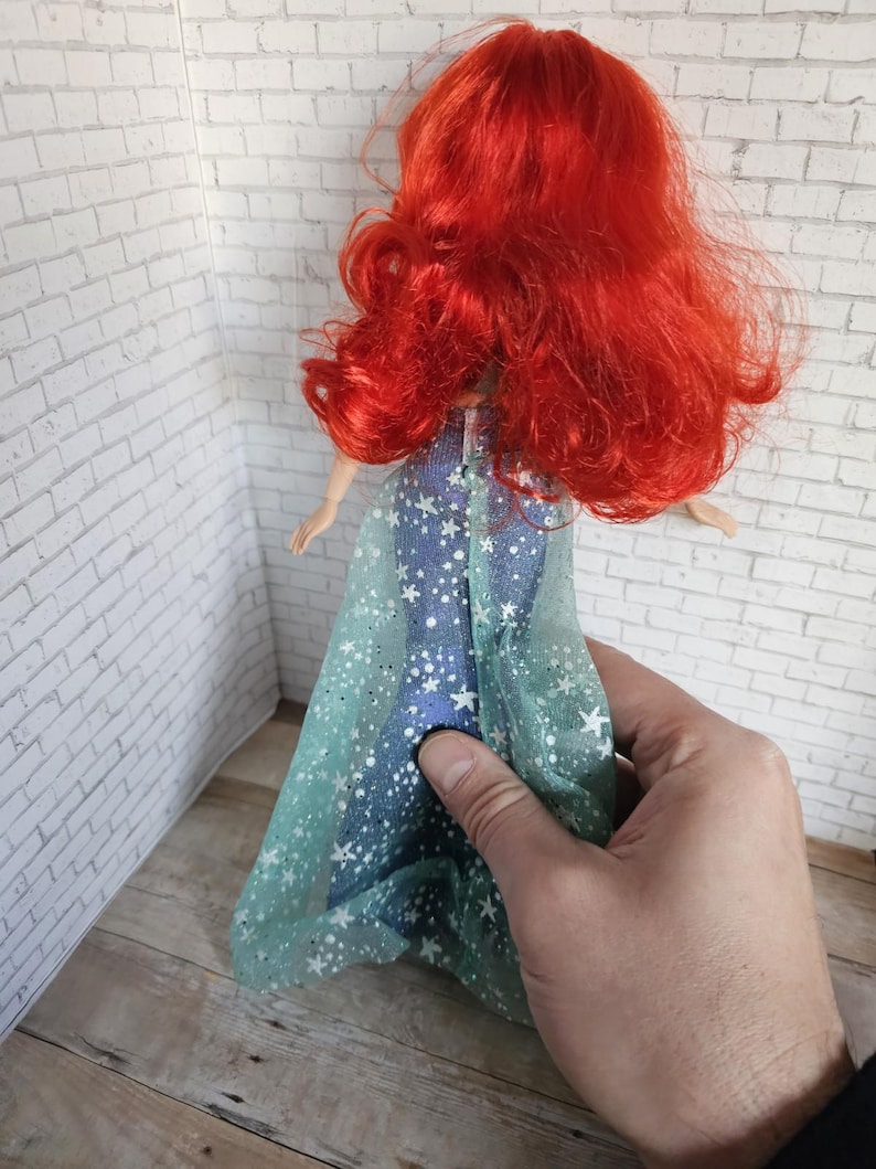 May include: A doll with bright red hair and a blue dress decorated with white stars. The doll's dress has a sheer, light blue overlay with white star accents. The doll is being held by a person.