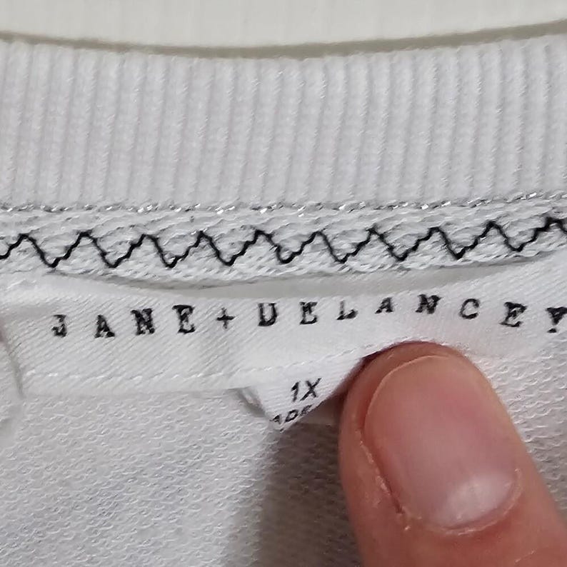 May include: Close-up of a white garment tag with the brand name "JANE + DELANCEY" in black capital letters. The tag also indicates a size of "1X". The garment has a silver and black trim.
