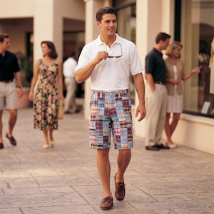 May include: A man wearing a white polo shirt and patchwork plaid shorts walks down a paved walkway. He holds sunglasses in his hand. Other people are in the background, some looking at a store window.