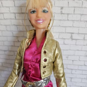 以下が含まれることがあります： 金色のジャケット、ピンクのトップス、シルバーのベルトを着た、ブロンドの髪の毛のドール。人形は緑色の目をし、微笑んでいます。背景は白いレンガの壁です。