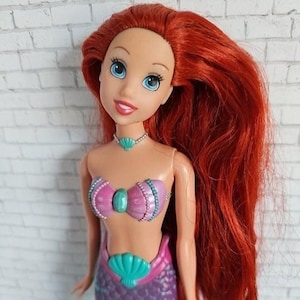 May include: A doll with long, vibrant red hair and blue eyes. The doll is wearing a pink and teal seashell-themed top and a purple mermaid tail. A teal seashell necklace completes the look. The doll is a popular character from a children's animated movie.