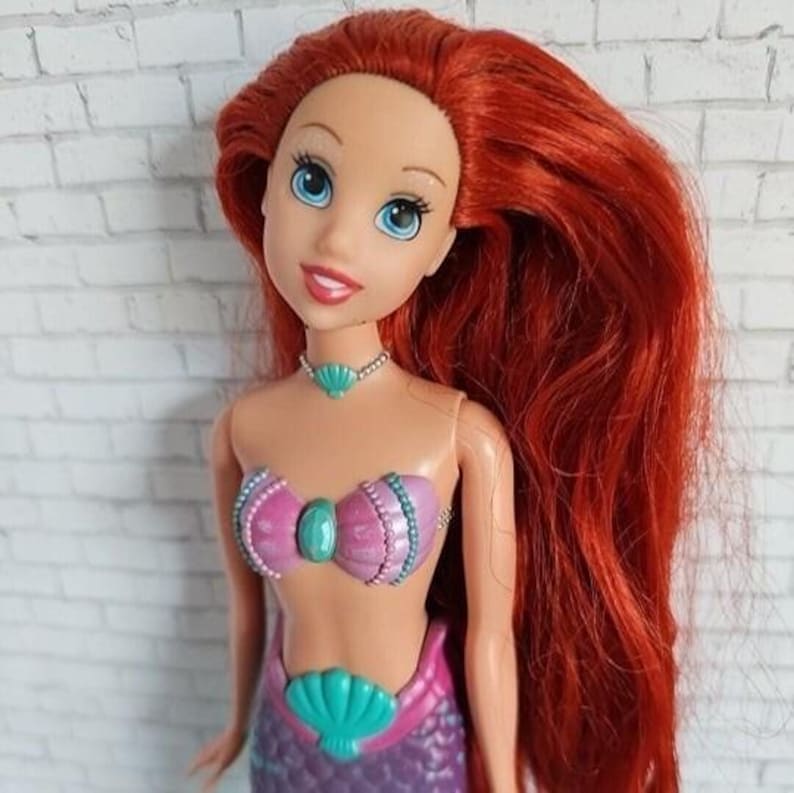 May include: A doll with long, vibrant red hair and blue eyes. The doll is wearing a pink and teal seashell-themed top and a purple mermaid tail. A teal seashell necklace completes the look. The doll is a popular character from a children's animated movie.