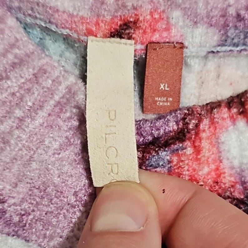 Puede incluir: Primer plano de una tela estampada floral en tonos rosa, morado y azul. La imagen incluye dos etiquetas: una con la marca "PILCR" y otra con "XL" y "MADE IN CHINA".