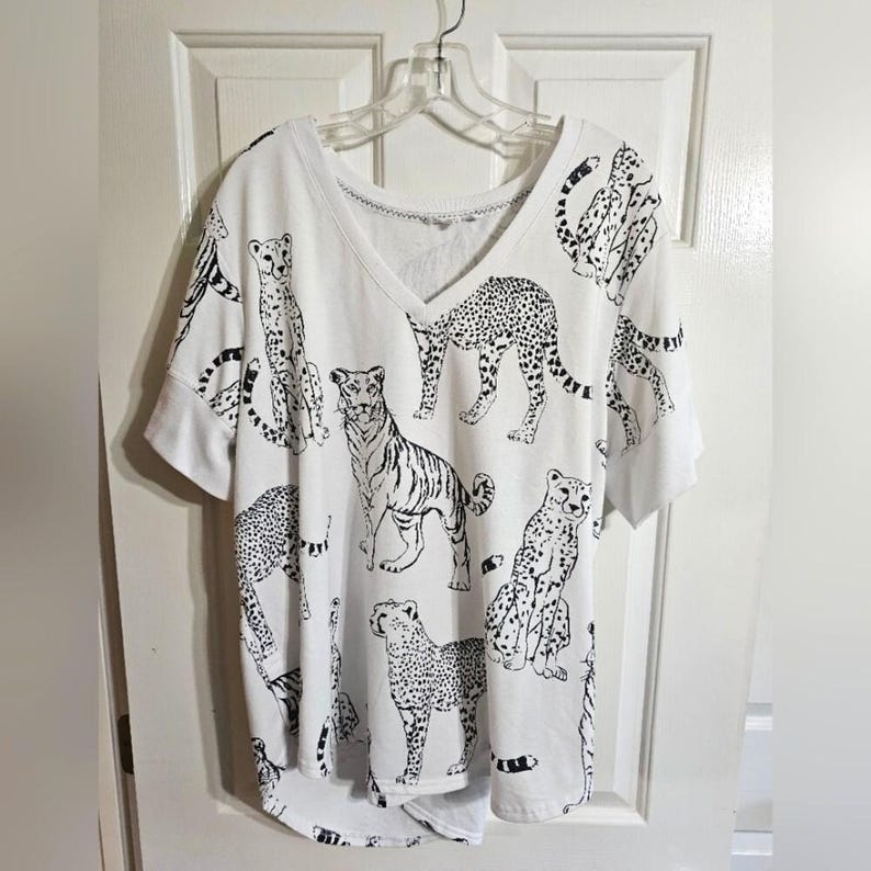 May include: White V-neck t-shirt with a black animal print. The print features various big cats, including leopards and tigers. The shirt has short sleeves and a relaxed fit, hanging on a clear plastic hanger.