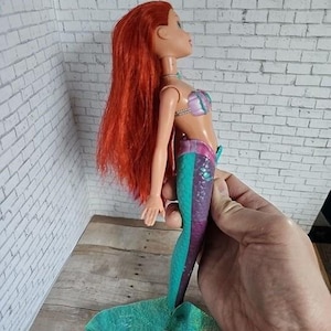 May include: A doll of a mermaid with long red hair, wearing a purple and pink seashell top and a teal and purple tail. The doll is held by a hand, showcasing the side profile of the toy.