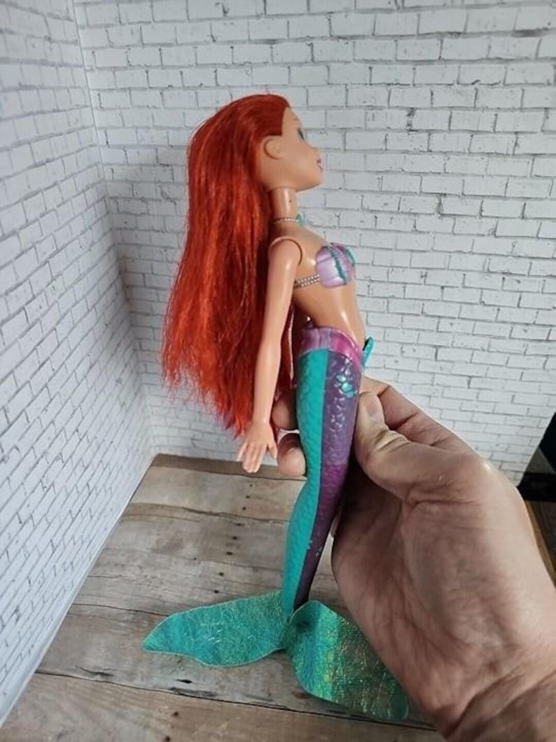 May include: A doll of a mermaid with long red hair, wearing a purple and pink seashell top and a teal and purple tail. The doll is held by a hand, showcasing the side profile of the toy.