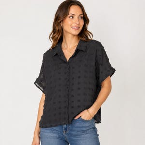 Puede incluir: Camisa negra de manga corta con un patrón de lunares texturizado. La camisa tiene cuello y se abrocha con botones. Se ven un collar y una pulsera dorados.