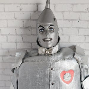 Könnte beinhalten: Eine silberfarbene Blechmann-Puppe mit spitzem Hut, blauen Augen und einer Fliege. Die Puppe hat eine herzförmige Uhr auf der Brust. Die Puppe ist eine Figur aus dem Zauberer von Oz.