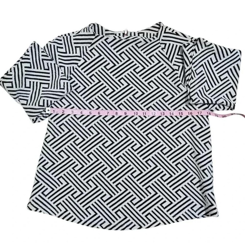 May include: A black and white geometric patterned top with short sleeves. The top is measured with a pink measuring tape, showing a length of approximately 18 inches.