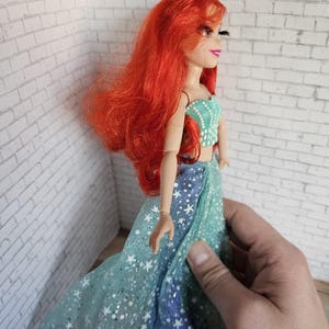 May include: A doll with long, vibrant red hair and a turquoise top with white accents. The doll wears a flowing, star-patterned gown in shades of blue and green. The doll is a collectible item.