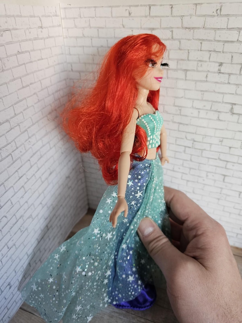 May include: A doll with long, vibrant red hair and a turquoise top with white accents. The doll wears a flowing, star-patterned gown in shades of blue and green. The doll is a collectible item.