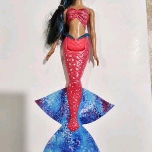 Puede incluir: Mu&ntilde;eca de sirena con top y cola rojos. La cola es roja con lunares blancos y hace la transici&oacute;n a una aleta estampada azul y blanca. La mu&ntilde;eca tiene pelo largo y negro y est&aacute; colocada sobre una superficie blanca.