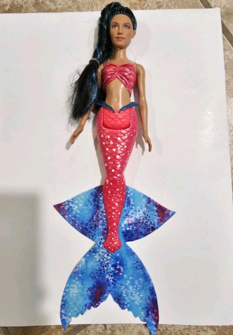 Puede incluir: Mu&ntilde;eca de sirena con top y cola rojos. La cola es roja con lunares blancos y hace la transici&oacute;n a una aleta estampada azul y blanca. La mu&ntilde;eca tiene pelo largo y negro y est&aacute; colocada sobre una superficie blanca.