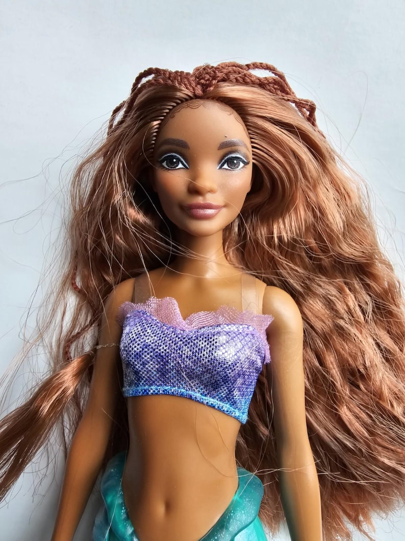 May include: A doll with brown skin, long auburn hair, and a purple and blue top. The doll has a smiling expression and is wearing a teal skirt. The doll's hair is styled with braids on top and flowing waves.