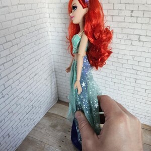 May include: A doll with long, bright red hair and a purple hairband. The doll is wearing a teal and blue dress with a starry pattern. The doll's skin is pale, and it is seen in profile.