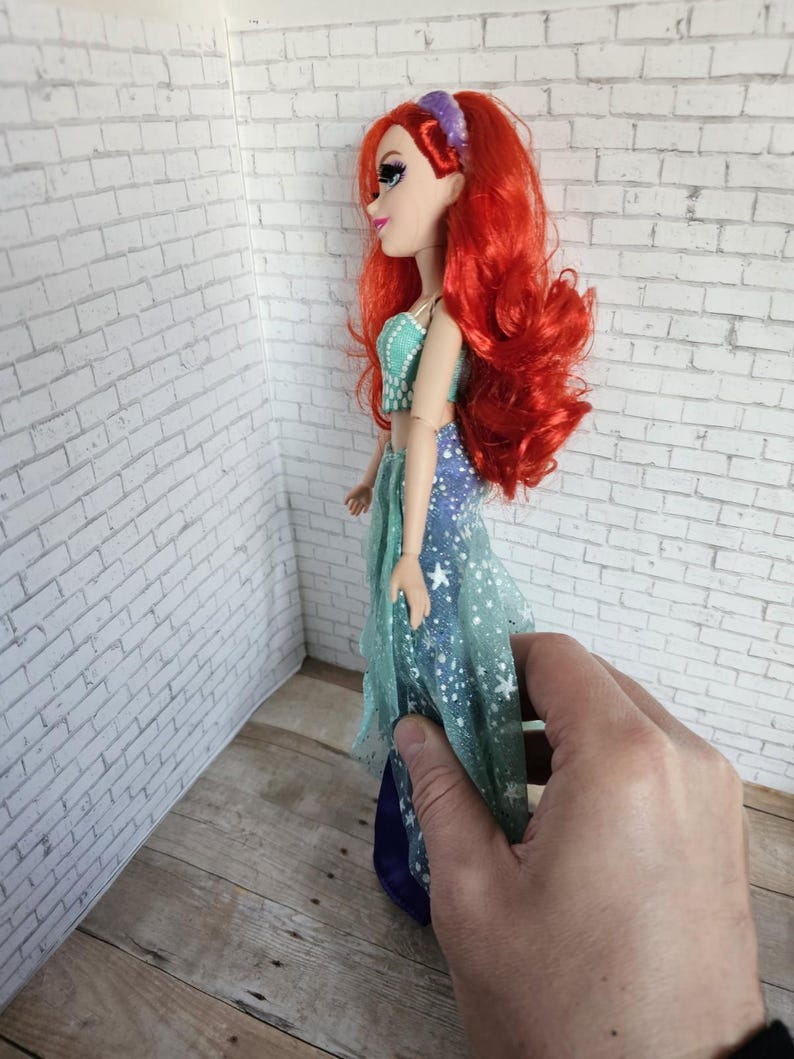 May include: A doll with long, bright red hair and a purple hairband. The doll is wearing a teal and blue dress with a starry pattern. The doll's skin is pale, and it is seen in profile.