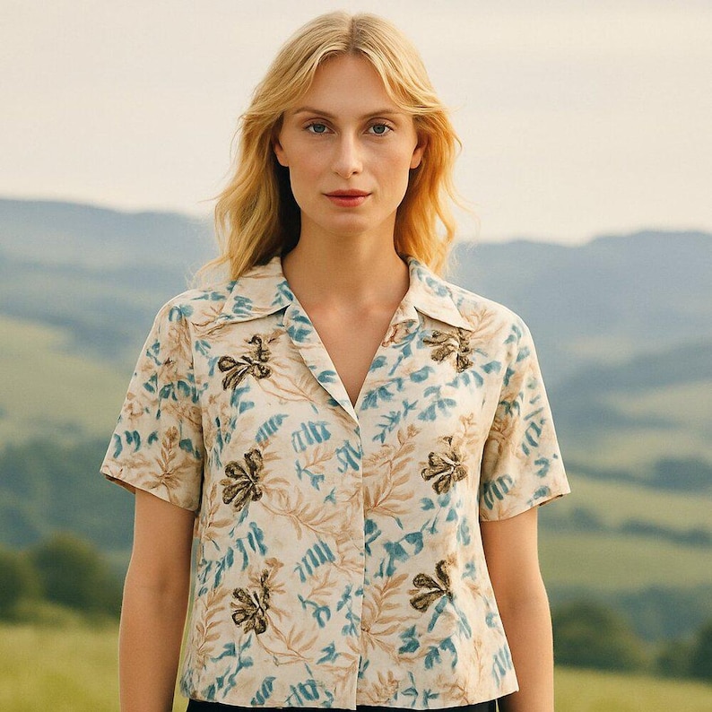 May include: A short-sleeved shirt with a floral pattern in beige, blue, and brown. The shirt has a collared neckline and button closure. The background shows a person in front of a blurred landscape.