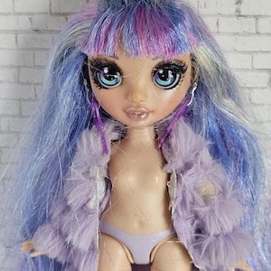 May include: A fashion doll with blue and purple hair, large blue eyes, and long eyelashes. The doll is wearing a lavender faux fur jacket and matching underwear. The doll has a fair complexion and is posed against a white brick wall.