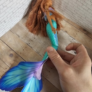 May include: A mermaid doll with long auburn hair, a blue and purple top, and a turquoise and blue tail. The doll's tail has large, fan-like fins in shades of blue and purple. The doll is held by a hand.