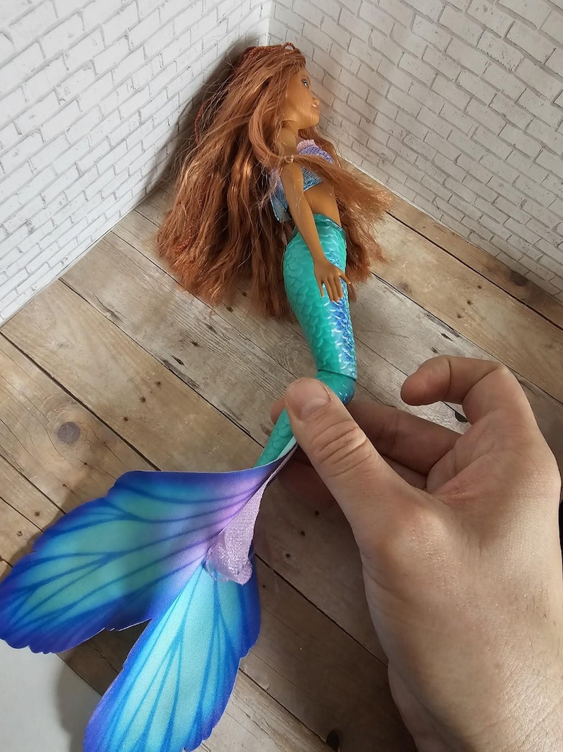 May include: A mermaid doll with long auburn hair, a blue and purple top, and a turquoise and blue tail. The doll's tail has large, fan-like fins in shades of blue and purple. The doll is held by a hand.