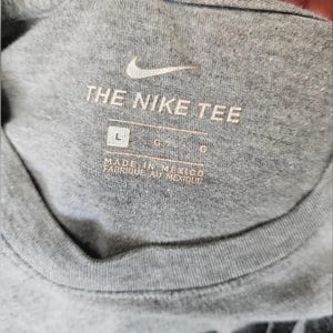 Pu&ograve; includere: Primo piano di un colletto di maglietta grigia con il logo Nike e la scritta "THE NIKE TEE". L'etichetta recita "MADE IN MEXICO FABRIQUE AU MEXIQUE" con indicatori di taglia.