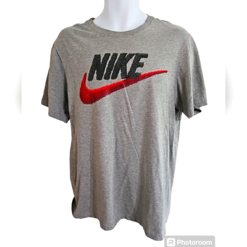 Pu&ograve; includere: T-shirt grigio a maniche corte con il logo Nike in nero e uno swoosh rosso. La maglietta &egrave; esposta su un manichino. Il logo Nike &egrave; ben visibile sulla parte anteriore della maglietta.