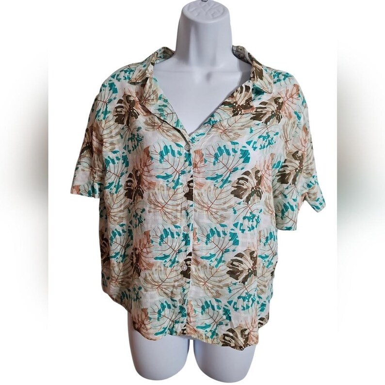May include: A short-sleeved button-down shirt with a tropical leaf pattern in shades of brown, teal, and peach on a white background. The shirt has a classic collar and is displayed on a white mannequin.