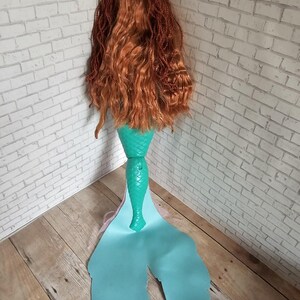 May include: A mermaid doll with long, auburn hair and a teal tail. The tail has a fin-like shape with a light blue gradient. The doll's hair is styled in a combination of braids and loose waves, and the tail is made of a shiny material.
