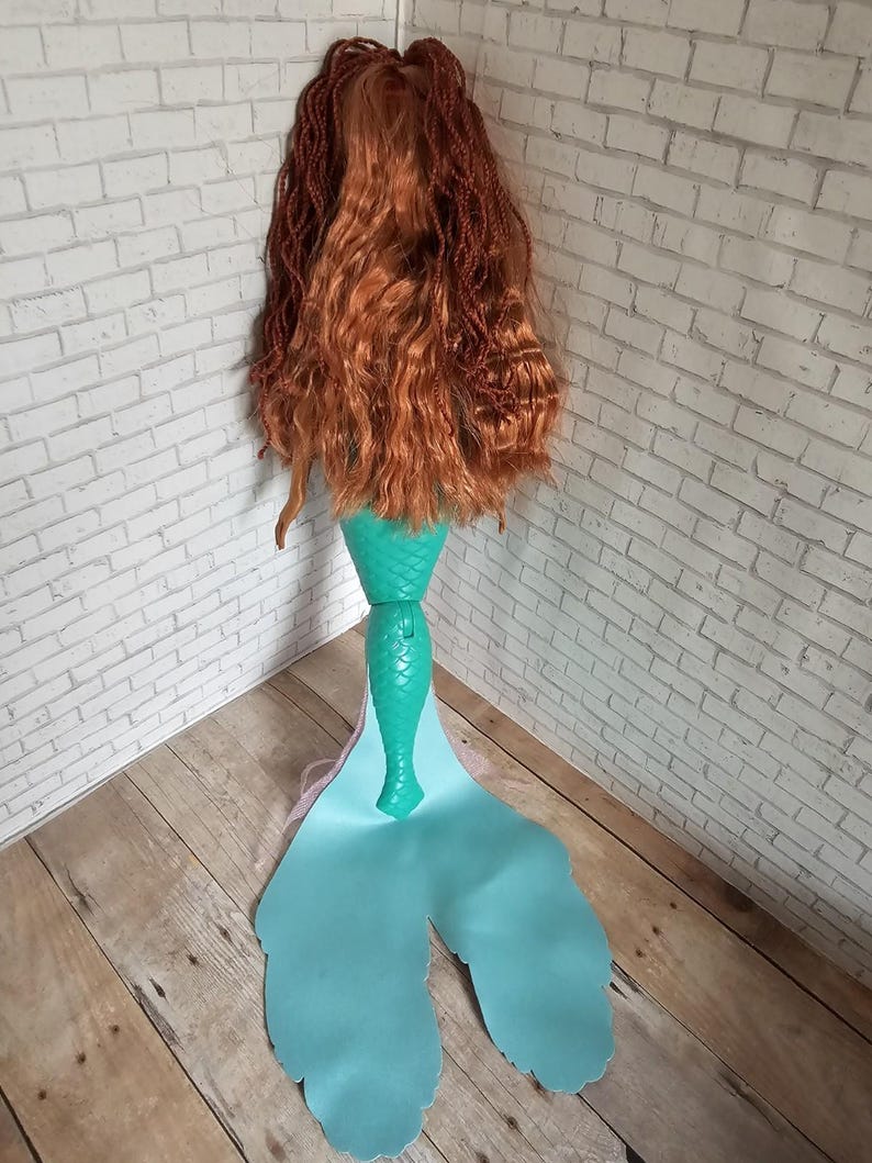 May include: A mermaid doll with long, auburn hair and a teal tail. The tail has a fin-like shape with a light blue gradient. The doll's hair is styled in a combination of braids and loose waves, and the tail is made of a shiny material.