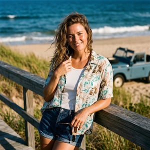 May include: A woman wearing a short-sleeved button-down shirt with a floral pattern in shades of brown, green, and blue. She is wearing a white tank top and denim shorts. The background includes a beach, ocean, and a light blue vehicle.