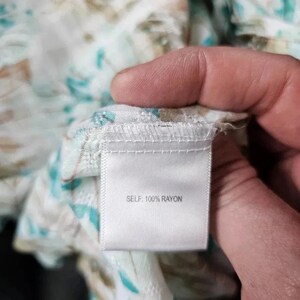 May include: Close-up of a white fabric tag with the text "SELF: 100% RAYON". The tag is attached to a floral patterned fabric with teal and brown accents. The fabric is held by a hand.