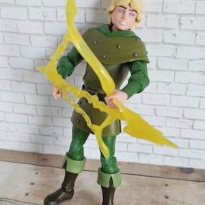 May include: A figurine of a character with blonde hair, wearing a green tunic, pants, and boots. The figure is holding a yellow, lightning-shaped bow. The background is a white brick wall.