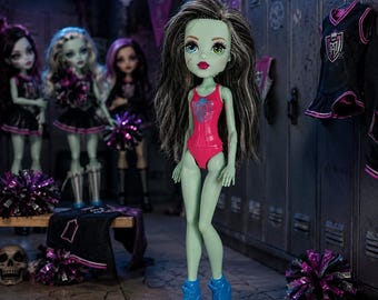 May include: A doll with light green skin, long dark hair with grey streaks, and bright red lips, wearing a pink leotard with a blue emblem and blue trainers. The doll stands in front of metal lockers with other dolls and purple pom-poms.