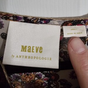 Pu&ograve; includere: Primo piano di un'etichetta di abbigliamento con il marchio "Maeve by Anthropologie" in lettere dorate su un'etichetta bianca. L'etichetta indica anche la taglia "Small" e "Made in India". Il tessuto ha un motivo floreale.