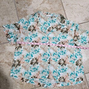 May include: A short-sleeved button-down shirt with a tropical leaf pattern in shades of brown, teal, and pink on a white background. The shirt is measured at approximately 23 inches across the chest.